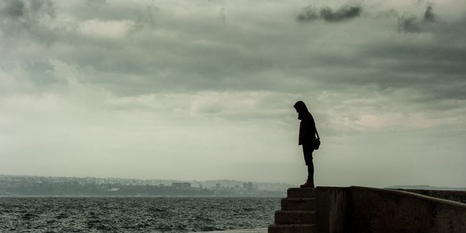 A silhouette of a person stands on a concrete structure by the sea under an overcast, cloudy sky.