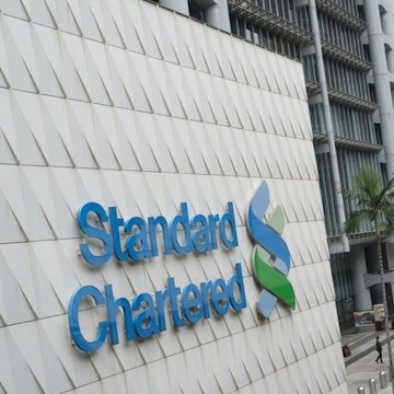 The image shows the blue "Standard Chartered" logo and name mounted on a textured white building facade, representing the banking partner mentioned in the post regarding the HKDAP stablecoin initiative. This ties directly to the announcement of Standard Chartered's collaboration with Animoca Brands on the Moca Chain, which the post anticipates will see positive movement this month.