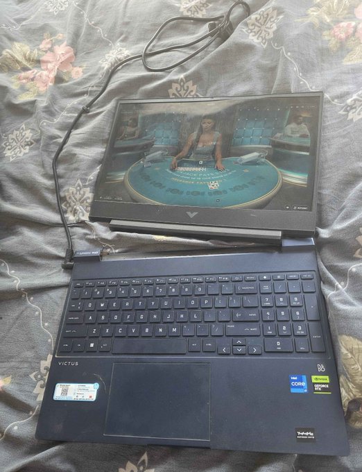 A dark blue HP Victus laptop is open on a bed, displaying a video game scene on its screen, with a charging cable connected.