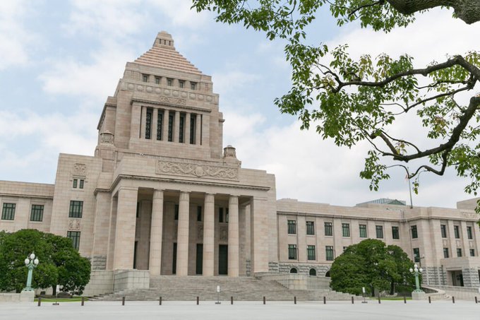 A daytime photograph of the National Diet Building of Japan, a grand stone government structure with columns and a central tower, framed by tree branches.