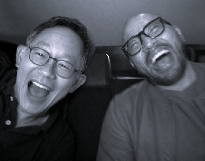 A black and white photo of two men with glasses laughing hysterically while sitting on a leather couch.