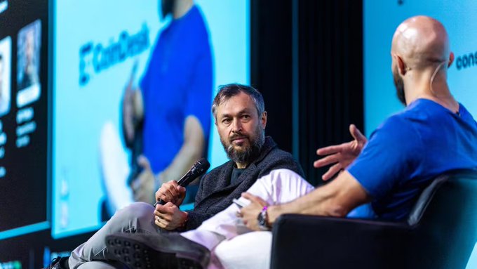 Two men sitting on chairs having a discussion on a stage with a 'CoinDesk' branded backdrop, one holding a microphone.