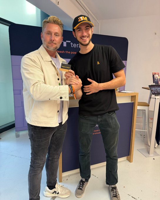 Two men stand together at what appears to be a crypto/NFT booth, with one holding a 'Zeneca Home Edition' NFT card; the younger man wears a 'ZM' cap and black t-shirt.