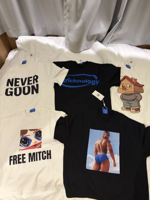 A flat lay of five T-shirts on a bed featuring designs including 'NEVER GOON', 'Kricknology', a cartoon house character, 'FREE MITCH' with a face, and a muscular man in blue trunks.