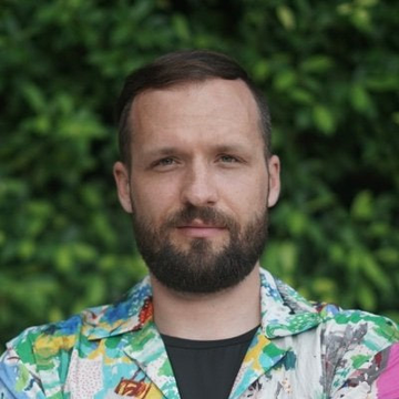 A man with a beard wearing a colorful patterned jacket over a dark shirt stands in front of green foliage. The image relates to the post about 1inch co-founder Anton Bukov announcing his departure from the company and his plans for a new venture called Second Tier.