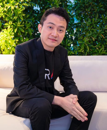A man wearing a black blazer over a black t-shirt with 'R' logo, sitting on a white couch with his hands clasped, against a backdrop of green plants.
