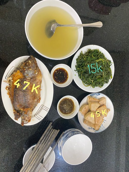 A meal laid out on a dark table featuring a fried fish, a bowl of broth, green vegetables, tofu, and dipping sauces, with prices written in yellow: 47K, 15K, and 45K.