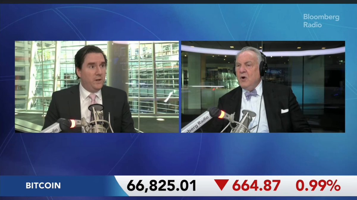 The image shows a news segment with two men talking into microphones, one wearing a suit and tie while the other has headphones around his neck. The background features a blue screen displaying the text "Charles Schwab Director of Research explains who is selling bitcoin". This suggests that Charles Schwab, the director of research at Schwab Capital Management, is discussing the topic of Bitcoin in this news segment.