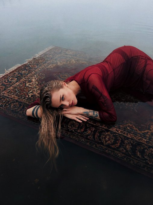 The image features a woman wearing a red dress, lying on her stomach with her head resting on a rug that appears to be floating in water. The background of the image is dark, which contrasts with the bright colors of the woman's dress and the rug she is laying on. The caption at the bottom of the photo reads "good morning photo by me + ai".