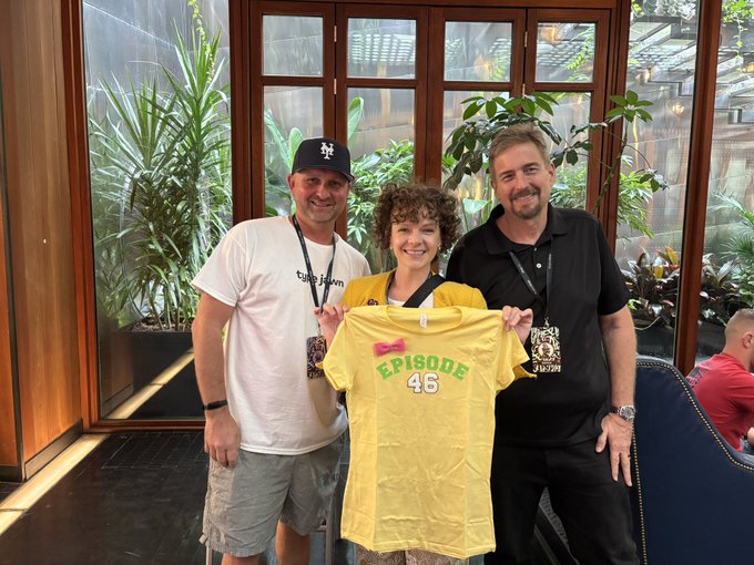 Three people pose together indoors holding a yellow t-shirt with "EPISODE 46" printed on it; a man wearing an LA Dodgers cap and "tulo fam" shirt, a woman in the middle, and a bearded man in a black polo shirt.