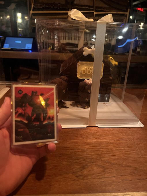 A hand holding a foil-stamped trading card in front of a clear display case containing a LEGO-style Domo-kun figure with bone horns, in what appears to be a café or shop.