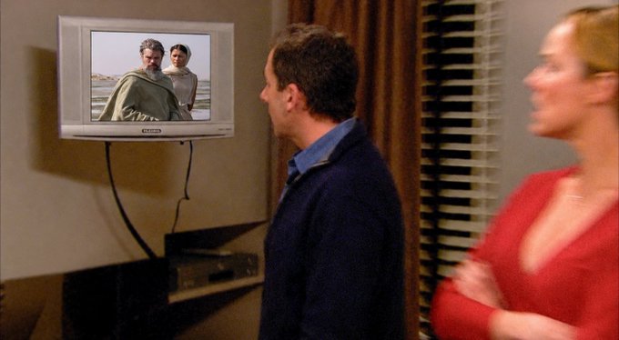 A man and woman stand watching a TV displaying an image of two people outdoors, appearing to be a scene from a film or TV show.