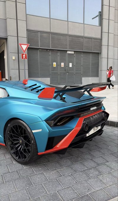 A blue and red Lamborghini Huracán STO supercar parked on a cobblestone street in front of a modern building, with a person walking in the background.