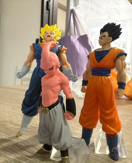 The image features three figurines of characters from the anime Dragon Ball Z, standing next to each other on a table. The figurines are dressed in different outfits, with one being pink and another orange, while the third one has an orange suit. They appear to be posed for a photo or displayed as collectibles.