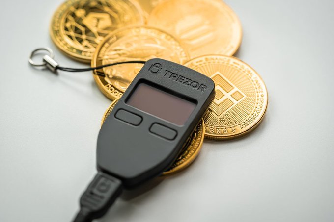 A black Trezor hardware wallet resting on top of several gold-colored cryptocurrency coins on a light gray surface.