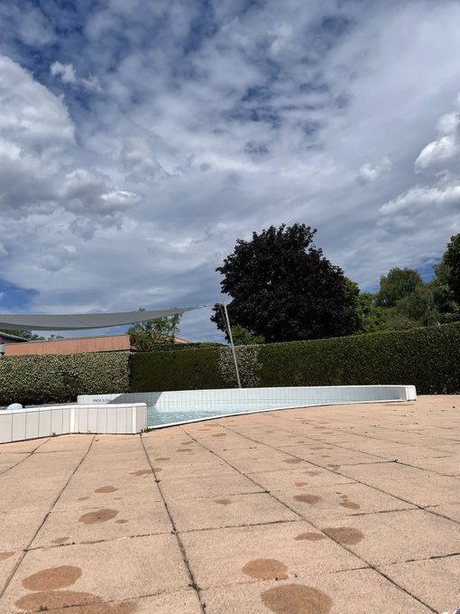 An outdoor scene of an empty swimming pool surrounded by tiled pavement, with green hedges, trees, and a dramatic cloudy blue sky in the background.