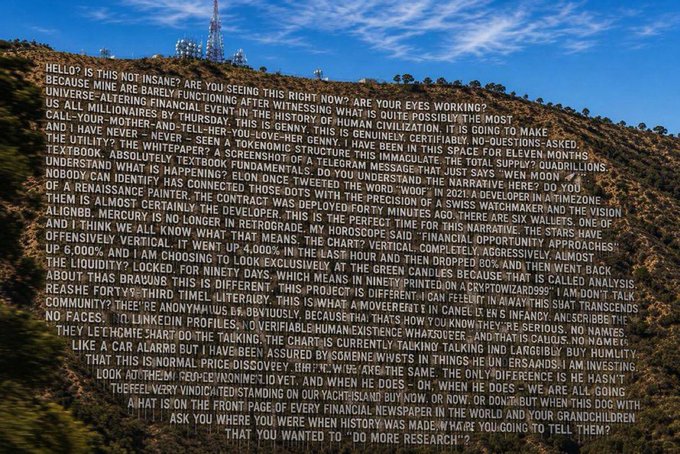 A hillside in San Francisco displays a large white text mural resembling a long string of gibberish or nonsense phrases, including "$HELLO??? IS THIS NOT INSANE???" at the beginning, with a communications tower visible atop the hill in the background.