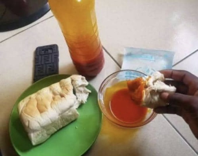A casual meal scene with a hand dipping bread into a bowl of red sauce on a tiled table, alongside a green plate with more bread, a plastic bottle of orange drink, a phone, and a face mask.