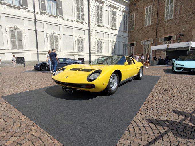 A bright yellow classic Lamborghini Miura sports car displayed on a dark mat at an outdoor car show in a cobblestone courtyard, with a man in a blue shirt standing beside it and other luxury vehicles visible in the background.