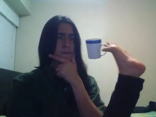 A person with long dark hair in a green jacket holds a blue and white mug thoughtfully near their face in a dimly lit room.