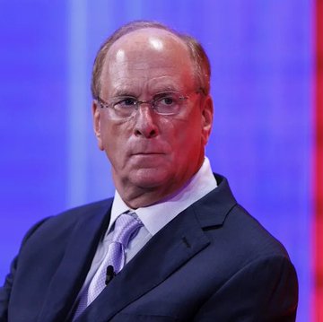 A man in a dark suit, light purple tie, and glasses appears against a blue and red background, likely in a news or interview setting.