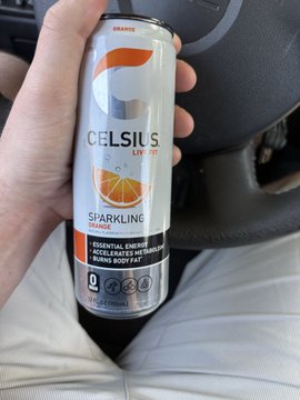 A hand holding a silver and orange 'Celsius Sparkling Orange' essential energy can inside a car.