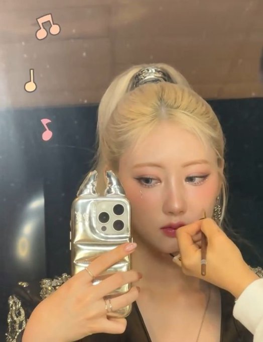 A blonde-haired woman takes a mirror selfie with a silver phone case, showcasing bold makeup with pink blush and graphic eyeliner, styled with her hair in a high ponytail. Decorative music note stickers float in the corner of the frame, complementing the artistic, performance-ready look associated with the artist SHIN RAMI.