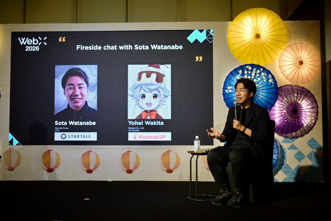 A man speaks into a microphone at the WebX 2026 'Fireside chat with Sota Watanabe', with a large screen showing his photo alongside a digital avatar labeled 'Yohei Wakita' from KudasaiJP, and colorful traditional Japanese parasols decorating the stage.