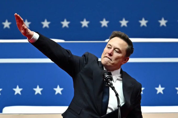Elon Musk stands at a podium, his arm raised in a dramatic gesture, with a backdrop featuring white stars on a blue field reminiscent of the American flag.