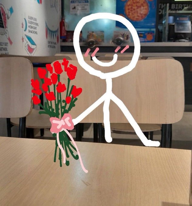 A simple white stick figure drawing of a smiling person holding a bouquet of red roses, overlaid on a photo of a wooden table in what appears to be a cafeteria.