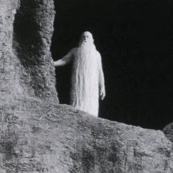 A grainy black-and-white photograph of a ghostly figure draped in white robes emerging from a dark doorway or cave-like opening.