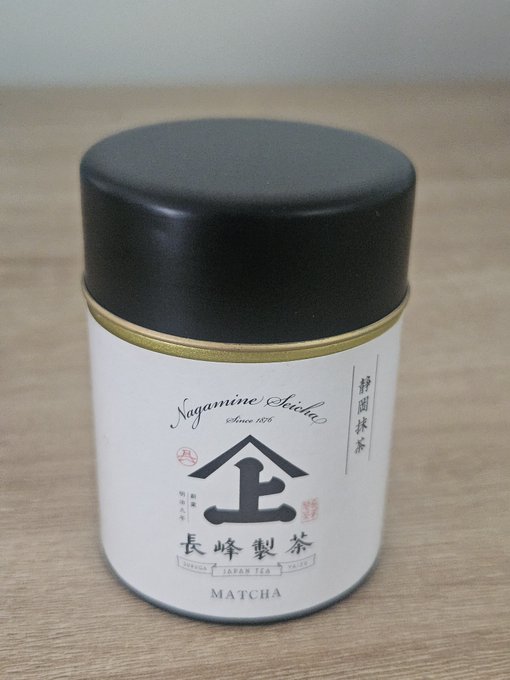 A white cylindrical tin can of 'SAGAMINE IRIICHA' Japanese green tea labeled 'MATCHA' with Japanese characters and a black lid sits on a wooden surface.