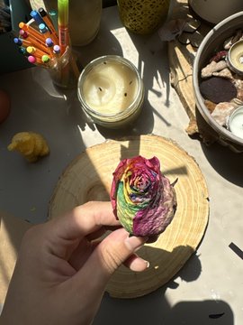 A hand holds a multicolored, rose-shaped object above a wooden surface, with a jar of white paste, a cup of colored pencils, and other craft items nearby.