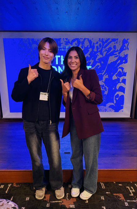 Two people stand on a stage pointing thumbs-up toward the camera, with one wearing a person identifier badge and a blue presentation screen behind them.