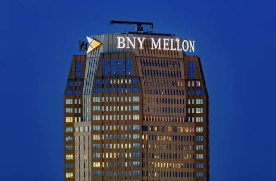 A skyscraper displays an illuminated "BNY MELLON" sign at its crown against a dark blue evening sky, with lit office windows visible across the building's facade. The image visually anchors the post's announcement of BNY Mellon's institutional adoption of blockchain technology to handle its massive $8.6 trillion in assets.