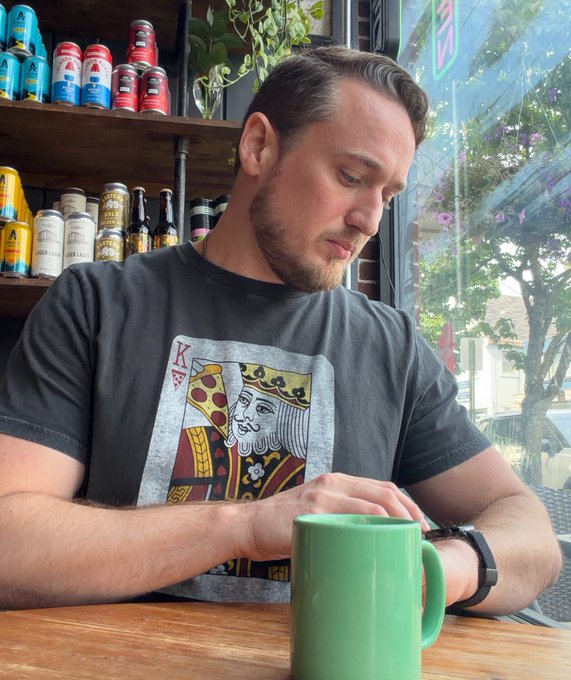 A bearded man wearing a black graphic t-shirt featuring a King of Diamonds design sits at a wooden table with a green ceramic mug, with shelves of colorful beverages visible behind him.