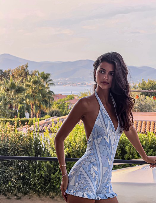 A brunette woman poses in a blue and white geometric-patterned halter dress, accessorized with MESHKI style and APM jewelry, against a scenic backdrop of palm trees, rolling hills, and a distant coastline at sunset.