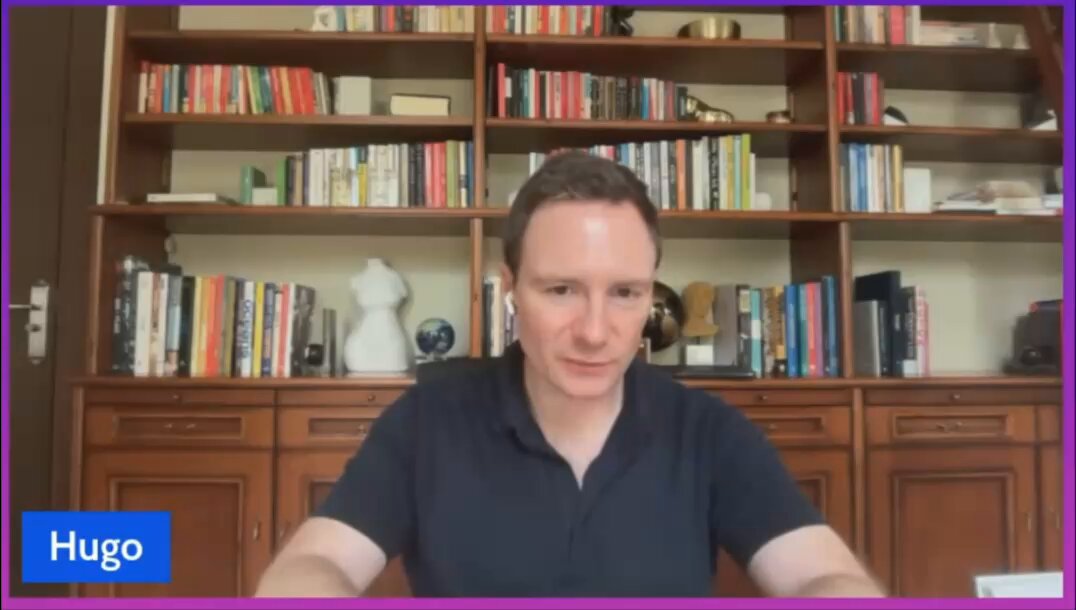 In the image, a man wearing a black shirt is sitting at a desk with his hands resting on the table. He appears to be looking directly into the camera while talking. The room has several bookshelves filled with various books of different sizes, indicating that this could be an office or study area. A computer monitor and keyboard are also present in front of him, suggesting that he might be working or studying from home.