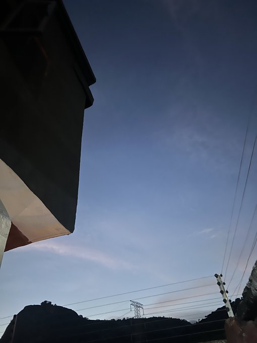 A photo looking up at a dusky blue sky with wispy clouds, showing the corner of a building roof on the left and power lines stretching across the frame above distant mountains.