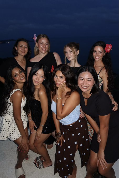 A group of eight young women pose together outdoors at night, with one in a white top and brown polka-dot pants in the center.