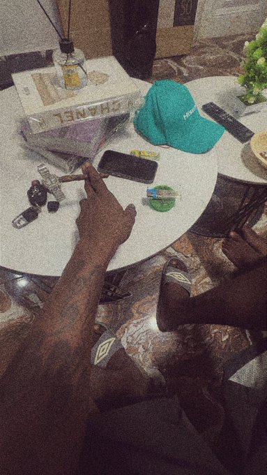 A first-person, grainy-filtered photo looking down at a hand reaching for a smartphone on a white round table, alongside a green cap, remote, and a box labeled 'CHANEL'.
