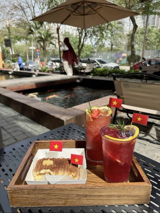 A wooden tray on an outdoor cafe table holding two glasses of red beverages garnished with lemon and a pastry, each marked with small Vietnamese flag toothpicks, with a koi pond in the background.