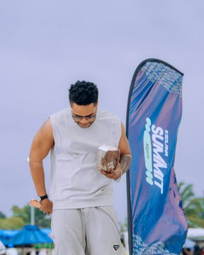 A man in a white sleeveless shirt and glasses smiles while holding a packaged item at an outdoor beach event, with a banner reading "SUNWAT" partially visible beside him, reflecting the fun and networking atmosphere of the Solana Summit Nigeria 2026 closing mixer hosted in Uyo.