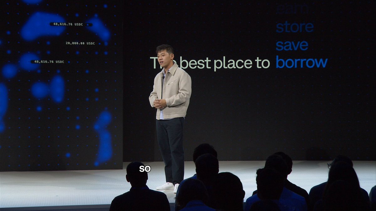 The image shows a man standing at a podium, giving a speech or presentation to an audience of people sitting behind him. The speaker appears to be addressing the crowd about the importance of keeping their Coinbase experience seamless and secure.