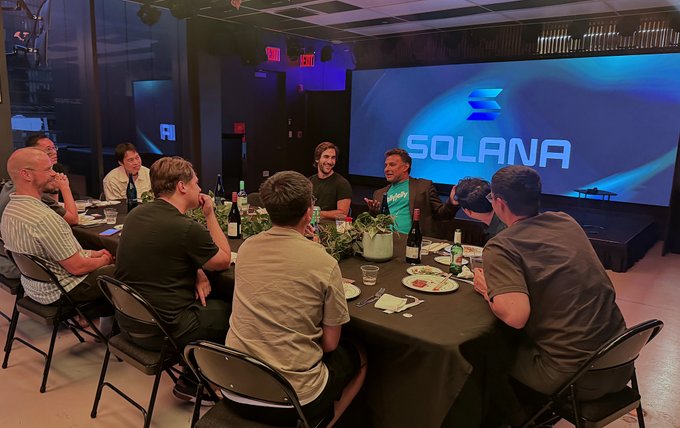 A group of people seated around a dinner table in front of a large screen displaying the SOLANA logo.