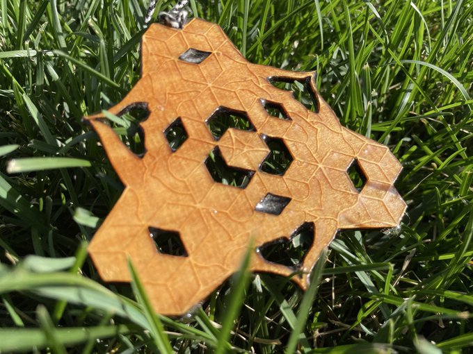 A close-up of an ornate, gold-colored leaf-shaped pendant or charm with geometric cutouts, lying on green grass.