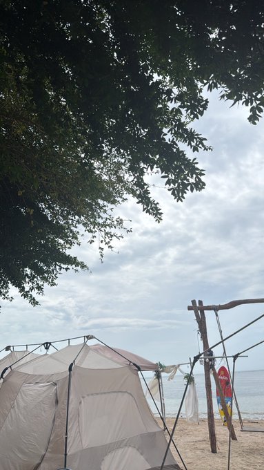 A beachside camping setup with a tent and a small red flag sits under a cloudy sky framed by tree branches, matching the relaxed weekend vibe of enjoying coffee on a Sunday morning getaway.