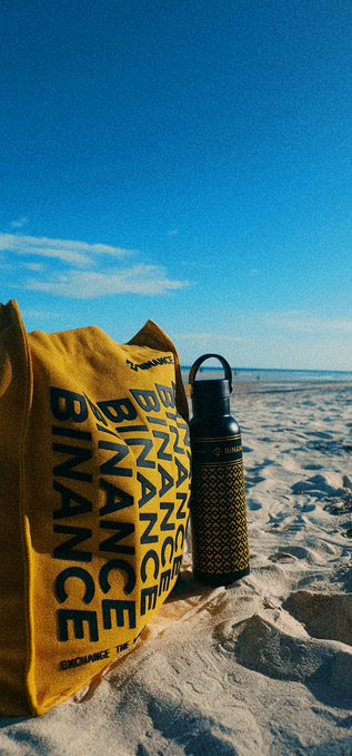 A yellow Binance tote bag and a matching patterned water bottle rest on a sandy beach, with the bold "BINANCE" branding clearly visible on both items under a bright blue sky—perfectly tying into a cheerful "GM" greeting promoting the crypto exchange.