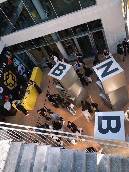 An overhead shot captures a crowd of people gathered outside a glass-fronted building near large cube sculptures spelling 'BNB' in black and white.