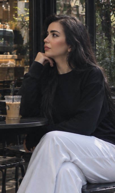 A woman with long dark hair sits thoughtfully at an outdoor café table, wearing a black sweater and white pants, with a coffee cup beside her.